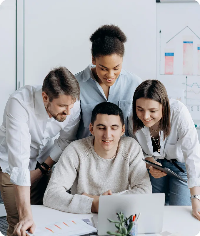 Four colleagues gathered around a laptop, looking engaged and smiling during a discussion in a bright office.